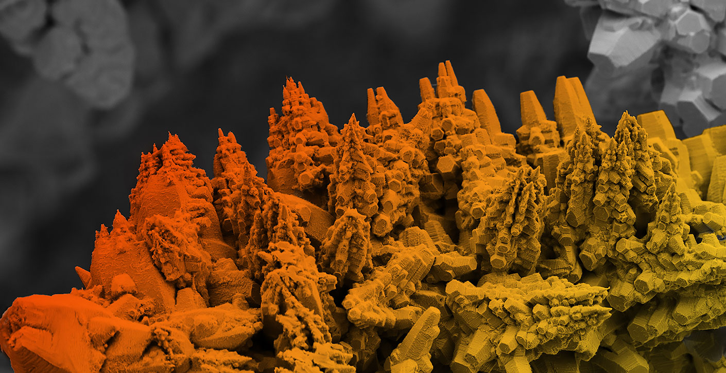 Image of steel topography in a scanning electron microscope, showing orange and yellow colored mountain-like structures against a grey background – visualizes internal material structures and possible inclusions. Image of steel topography in a scanning electron microscope, showing orange and yellow colored mountain-like structures against a grey background – visualizes internal material structures and possible inclusions.
