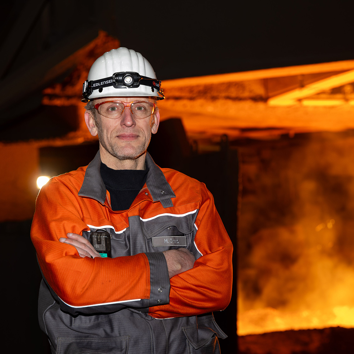 Marc Orenz, wearing orange-grey protective clothing and a white helmet, stands with his arms crossed in front of a glowing industrial facility in a steel mill.
