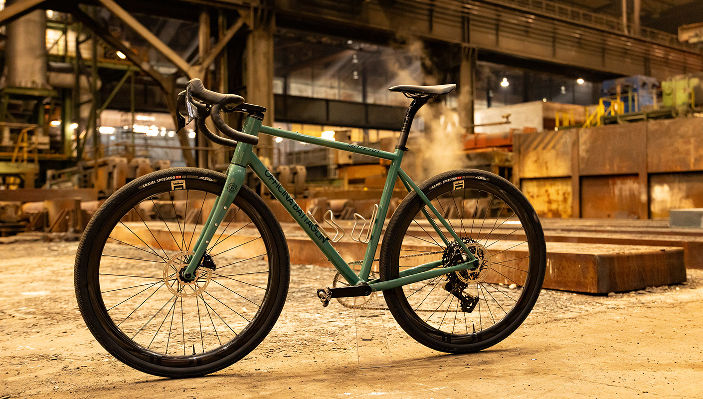 A steel bicycle stands in an industrial hall of a steel mill, positioned in front of production equipment and steel slabs.
