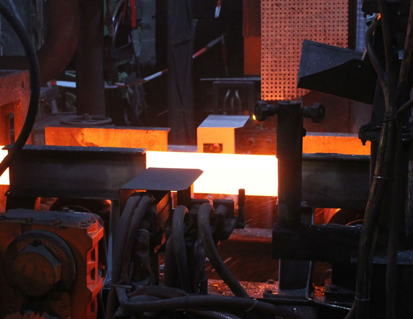 A glowing hot bloom moves along the roller table towards the cooling bed. The workpiece glows orange-red against a dark background. Two cameras above monitor the surface of the bloom for material defects. A glowing hot bloom moves along the roller table towards the cooling bed. The workpiece glows orange-red against a dark background. Two cameras above monitor the surface of the bloom for material defects.