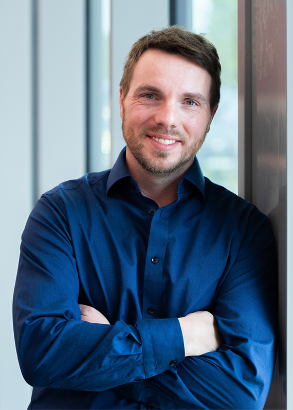 Christian Kühlen steht mit verschränkten Armen im blauen Hemd an ein Fenster gelehnt in einem modernen Bürogebäude.