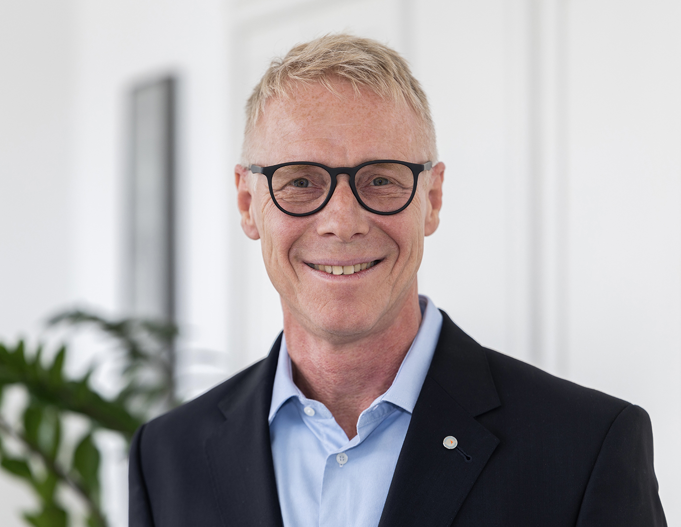 Portrait photo of Dr. Eng. Benedikt Ritterbach. He is wearing glasses, a white shirt, a dark tie, and a dark blazer with a lapel pin. He is smiling warmly at the camera against a warmly lit background with a color gradient.