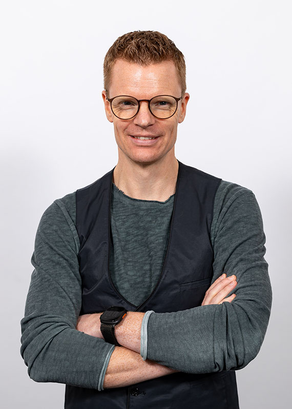 Portrait of Stefan Mecke. Portrait of Stefan Mecke. He is wearing a gray sweater and a black vest and is smiling at the camera.
