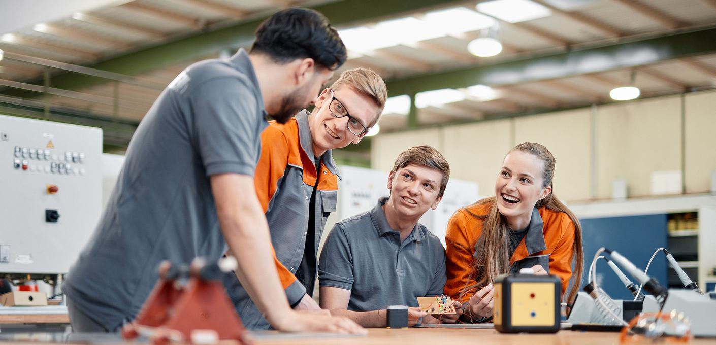 Junge Mitarbeitende an der Werkbank Vier junge Mitarbeitende in grauer und orangefarbener Arbeitskleidung stehen gemeinsam an einer Werkbank. Sie betrachten lachend ein Werkstück und wirken in ein Gespräch vertieft. Im Hintergrund sind Maschinen und Werkzeuge zu erkennen.