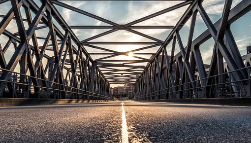 Stahlfachwerkbrücke Blick aus niedriger Kameraperspektive über eine Stahlfachwerkbrücke. Die symmetrische Konstruktion aus massiven Stahlträgern führt den Blick in die Tiefe, wo die untergehende Sonne durch das Fachwerk scheint. Die Mittelmarkierung der Fahrbahn leuchtet im warmen Gegenlicht. Am Horizont sind Gebäude erkennbar.