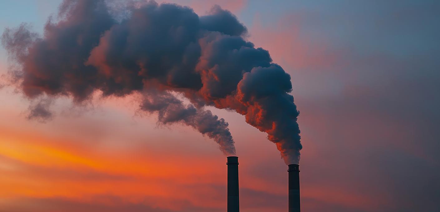 Industrieschornsteine stoßen Rauch aus Zwei Industrieschornsteine stoßen dichten Rauch vor einem rot-orangen Abendhimmel aus.