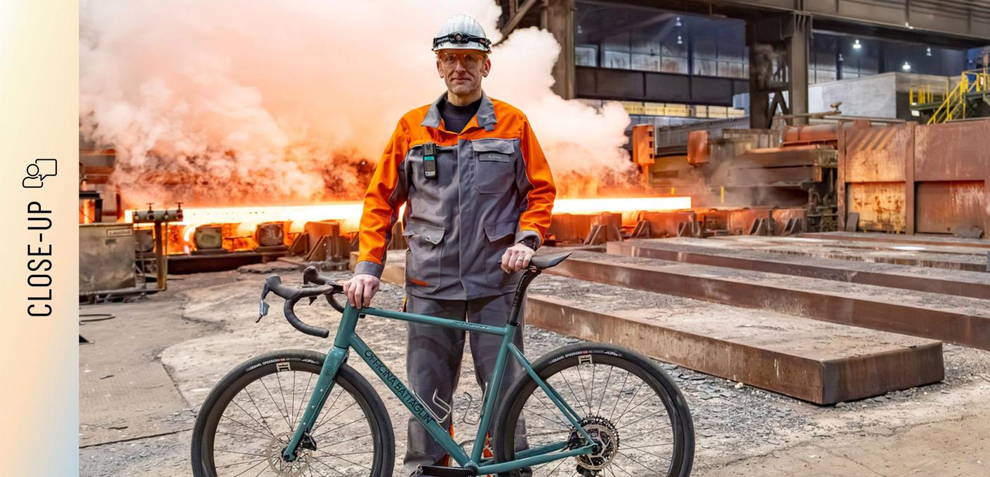 Marc Orenz, wearing orange-grey protective clothing and a helmet, stands in a steel mill holding a bicycle. In the background, glowing steel slabs and rising steam are visible.