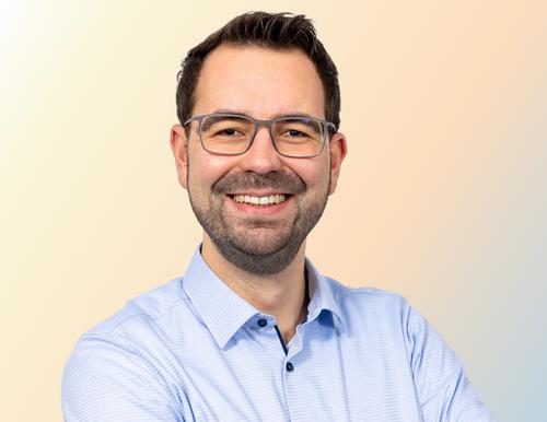 Portrait photo of Lennart Schümann, expert for energy strategy and procurement at Salzgitter AG. He wears a light blue shirt and glasses and smiles friendly into the camera against a light background.