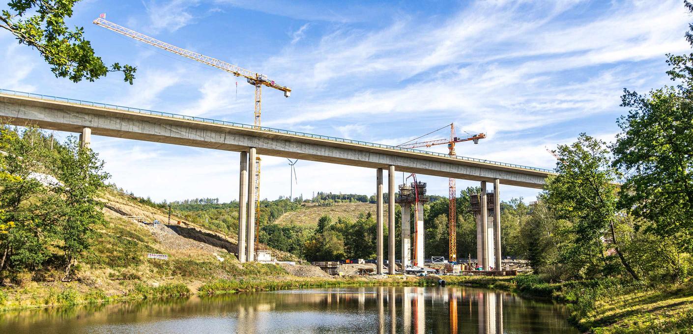 Baustelle einer Autobahnbrücke auf hohen Betonpfeilern mit zwei Baukränen, grüne Hügellandschaft im Hintergrund, Spiegelung der Konstruktion in einem Gewässer im Vordergrund. Baustelle einer Autobahnbrücke auf hohen Betonpfeilern mit zwei Baukränen, grüne Hügellandschaft im Hintergrund, Spiegelung der Konstruktion in einem Gewässer im Vordergrund.