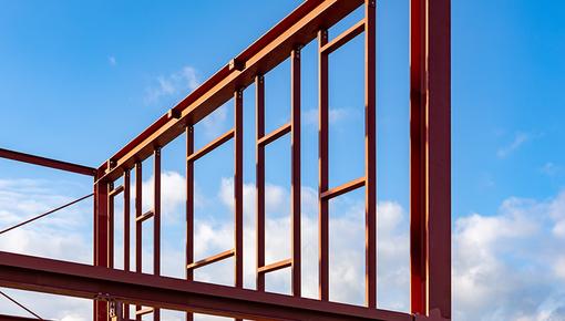 Rote Stahlskelettkonstruktion eines Wohngebäudes in der Bauphase vor blauem Himmel mit weißen Wolken – zeigt die tragende Struktur aus Stahlprofilen