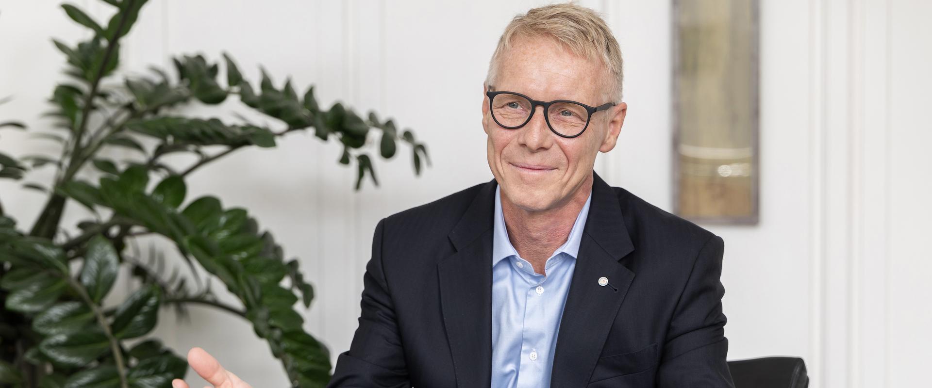 Porträtfoto von Benedikt Ritterbach, Geschäftsführer der Salzgitter Mannesmann Forschung GmbH (SZMF). Er trägt eine Brille, ein weißes Hemd und ein dunkles Sakko und blickt freundlich in die Kamera. Im Hintergrund sind Pflanzen und ein helles Interieur zu erkennen.