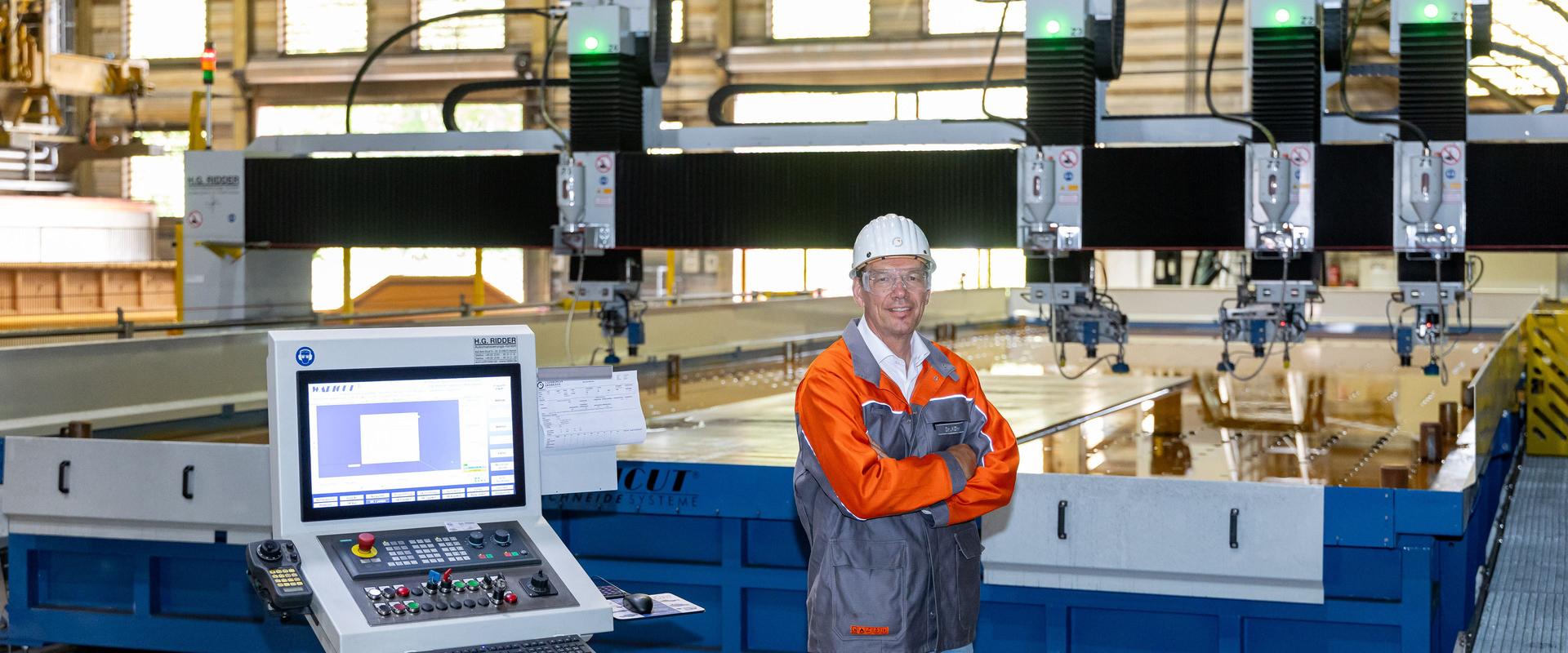 Portrait of Robert Kühn in front of a waterjet cutting machine Robert Kühn stands with his arms folded in a workshop in front of a large-format waterjet cutting machine. The control panel and screen can be seen in the foreground; several cutting heads of the machine are visible in the background.