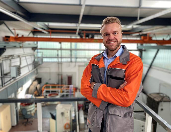 Porträtfoto von Alexander Gering, Gruppenleiter im Bereich Mechanische Prüfung bei der Salzgitter Mannesmann Forschung GmbH in Duisburg. Er trägt orangefarbene Arbeitskleidung und steht mit verschränkten Armen in einer Prüfhalle. Er lächelt freundlich in die Kamera. Im Hintergrund sind Industrieanlagen und Hallenstrukturen zu erkennen.