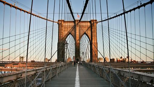 Bild der Brooklyn Bridge. Blick entlang des Fußgängerstegs der Brooklyn Bridge in Richtung eines der steinernen Pylontürme. Stahlseile spannen sich symmetrisch von beiden Seiten zur Mitte. Ein weißer Mittelstreifen führt den Blick auf den gotischen Spitzbogen des sandsteinfarbenen Turms zu, auf dem eine US-Flagge weht. Fußgänger sind in der Ferne erkennbar, das warme Licht der Abendsonne beleuchtet die Szene.