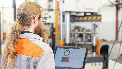 Porträtfoto von Fabian Spiegel, Data Scientist bei der Salzgitter Mannesmann Forschung GmbH. Er steht in grauer Arbeitskleidung mit orangefarbener Warnweste mit dem Rücken zur Kamera und blickt auf einen Bildschirm mit farbigen Diagrammen. Im Hintergrund ist eine industrielle Prüfanlage mit orangefarbenen Stützen zu erkennen.