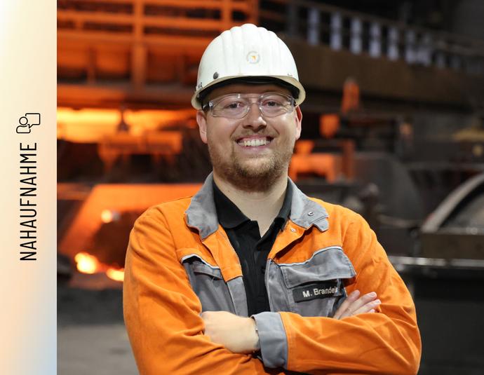 Marvin Brandeis steht mit verschränkten Armen und einem Lächeln vor einem Hochofen. Er trägt eine orangefarbene Arbeitsschutzjacke mit seinem Namenszug, einen weißen Schutzhelm und eine Schutzbrille. Im Hintergrund glüht das rötliche Licht der Produktionsanlage.