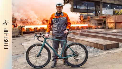 Marc Orenz with steel bicycle in steel mill Marc Orenz, wearing orange-grey protective clothing and a helmet, stands in a steel mill holding a bicycle. In the background, glowing steel slabs and rising steam are visible.