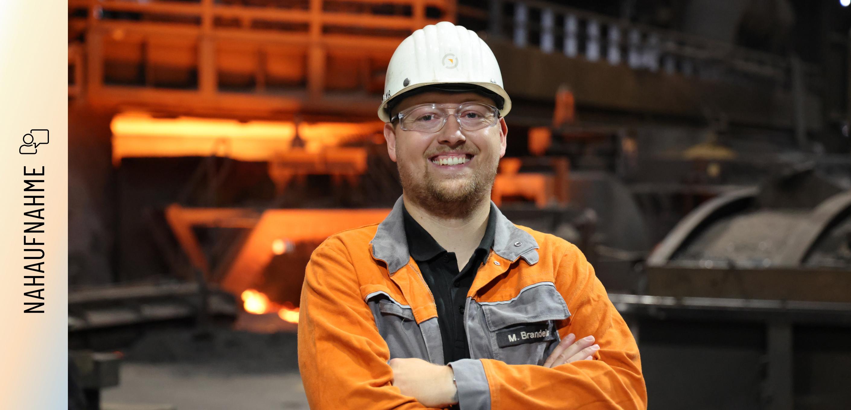 Marvin Brandeis steht mit verschränkten Armen und einem Lächeln vor einem Hochofen. Er trägt eine orangefarbene Arbeitsschutzjacke mit seinem Namenszug, einen weißen Schutzhelm und eine Schutzbrille. Im Hintergrund glüht das rötliche Licht der Produktionsanlage.