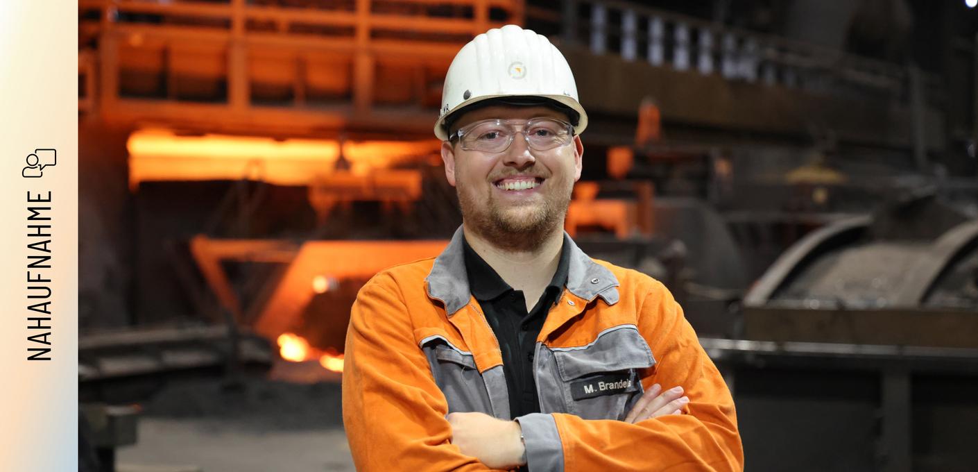 Porträt von Marvin Brandeis Marvin Brandeis steht mit verschränkten Armen und einem Lächeln vor einem Hochofen. Er trägt eine orangefarbene Arbeitsschutzjacke mit seinem Namenszug, einen weißen Schutzhelm und eine Schutzbrille. Im Hintergrund glüht das rötliche Licht der Produktionsanlage.