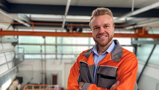 Porträtfoto von Alexander Gering, Gruppenleiter im Bereich Mechanische Prüfung bei der Salzgitter Mannesmann Forschung GmbH in Duisburg. Er trägt orangefarbene Arbeitskleidung und steht mit verschränkten Armen in einer Prüfhalle. Er lächelt freundlich in die Kamera. Im Hintergrund sind Industrieanlagen und Hallenstrukturen zu erkennen.