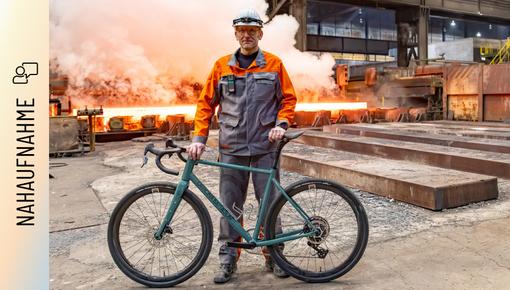 Marc Orenz in orange-grauer Schutzkleidung und Helm steht in einem Stahlwerk und hält ein Fahrrad, im Hintergrund glühende Stahlbrammen und aufsteigender Dampf.