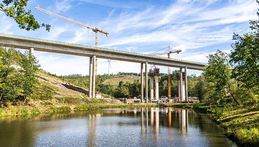 Baustelle einer Autobahnbrücke auf hohen Betonpfeilern mit zwei Baukränen, grüne Hügellandschaft im Hintergrund, Spiegelung der Konstruktion in einem Gewässer im Vordergrund. Baustelle einer Autobahnbrücke auf hohen Betonpfeilern mit zwei Baukränen, grüne Hügellandschaft im Hintergrund, Spiegelung der Konstruktion in einem Gewässer im Vordergrund.