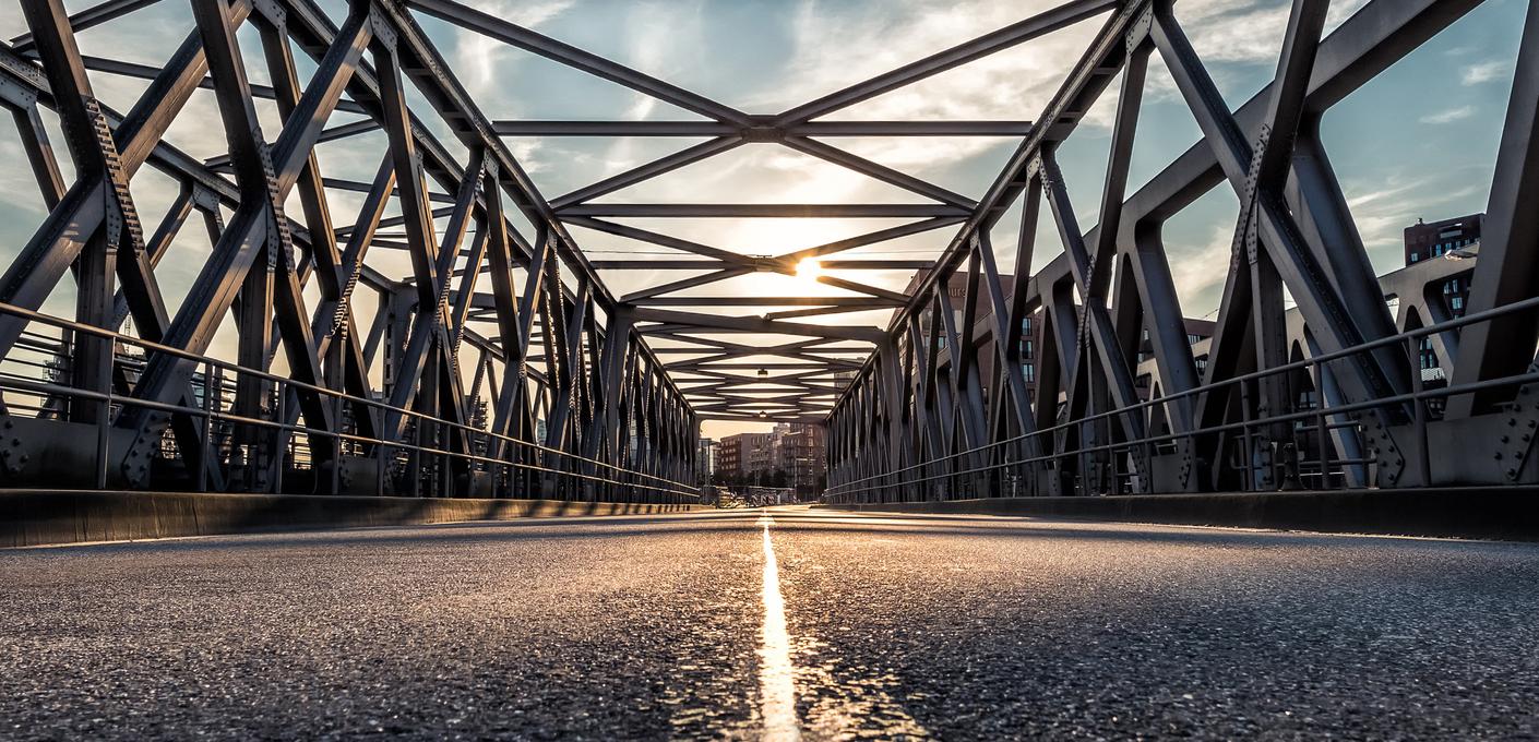Stahlfachwerkbrücke Blick aus niedriger Kameraperspektive über eine Stahlfachwerkbrücke. Die symmetrische Konstruktion aus massiven Stahlträgern führt den Blick in die Tiefe, wo die untergehende Sonne durch das Fachwerk scheint. Die Mittelmarkierung der Fahrbahn leuchtet im warmen Gegenlicht. Am Horizont sind Gebäude erkennbar.