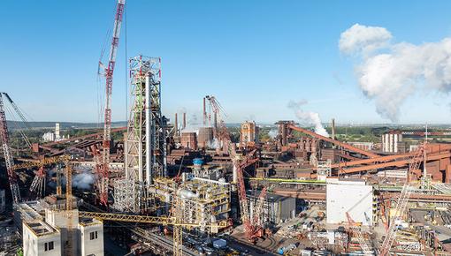 Industriegelände mit Kränen und Rohrleitungen Weitläufiges Industriegelände mit vielen Kränen, Rohrleitungen und rauchenden Schornsteinen an einem klaren Tag.
