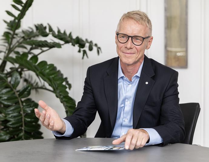 Portrait photo of Benedikt Ritterbach, Managing Director of Salzgitter Mannesmann Forschung GmbH (SZMF). He wears glasses, a white shirt and a dark blazer and looks friendly into the camera. Blurred plants and a bright interior are visible in the background.