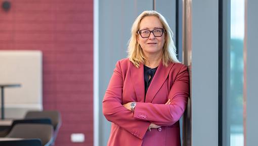 Ms. Potrafki is standing with her arms crossed, leaning against a window in a modern meeting room.