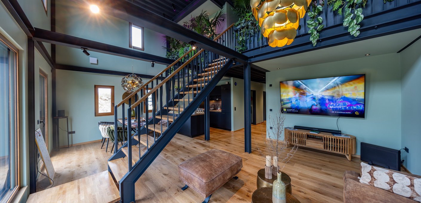 Offener Wohnbereich im Green Steel Home mit hoher Decke, sichtbarer schwarzer Stahlkonstruktion, moderner Holztreppe zur Galerie, hellblau gestrichenen Wänden, Holzboden, gelbem Designleuchtenpaar und Flachbildfernseher – zeigt flexible Raumgestaltung mit nachträglich versetzbaren Wänden.