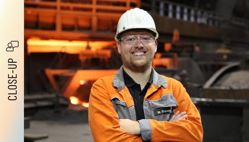Marvin Brandeis stands in front of a blast furnace with his arms crossed and a smile on his face. He is wearing an orange safety jacket with his name on it, a white hard hat, and safety goggles. In the background, the reddish light of the production facility glows.