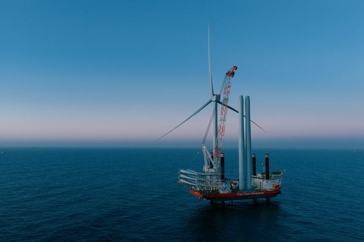 An installation vessel carrying components for an offshore wind turbine. An offshore installation vessel with a crane and wind turbine components on the open sea under a clear sky.