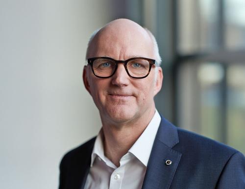 Sebastian Bross im Porträt Herr Bross im Porträt: Er trägt einen dunkelblauen Anzug und ein weißes Hemd und steht vor einem Fenster in einem modernen Gebäude.