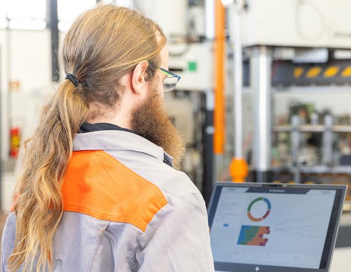 Porträtfoto von Fabian Spiegel, Data Scientist bei der Salzgitter Mannesmann Forschung GmbH. Er steht in grauer Arbeitskleidung mit orangefarbener Warnweste mit dem Rücken zur Kamera und blickt auf einen Bildschirm mit farbigen Diagrammen. Im Hintergrund ist eine industrielle Prüfanlage mit orangefarbenen Stützen zu erkennen.