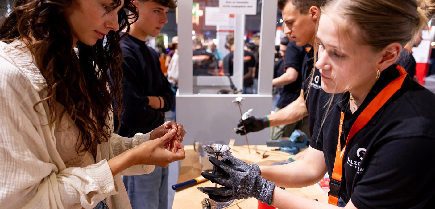A young female and a young male visitor craft steel flowers at a hands-on exhibit at the IdeenExpo. A female and a male employee in black clothing with Salzgitter branding assist them.