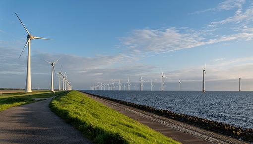 Offshore-Windpark Panoramablick auf einen Offshore-Windpark auf See. Mehrere große Windkraftanlagen ragen aus dem Wasser in einen leicht bewölkten Himmel. Im Vordergrund ist ein grüner Deich zu sehen, dahinter einige weitere Windkraftanlagen an Land.