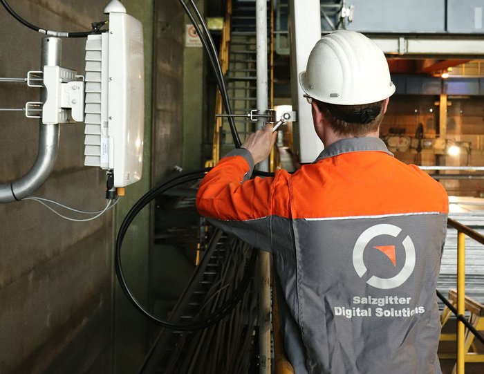 Ein Mitarbeiter in orangefarbener Arbeitskleidung mit dem Logo der Salzgitter Digital Solutions auf dem Rücken und weißem Schutzhelm montiert eine 5G-Funkeinheit an einer Wand in einer Industriehalle. Kabelstränge verlaufen unterhalb des Geräts.