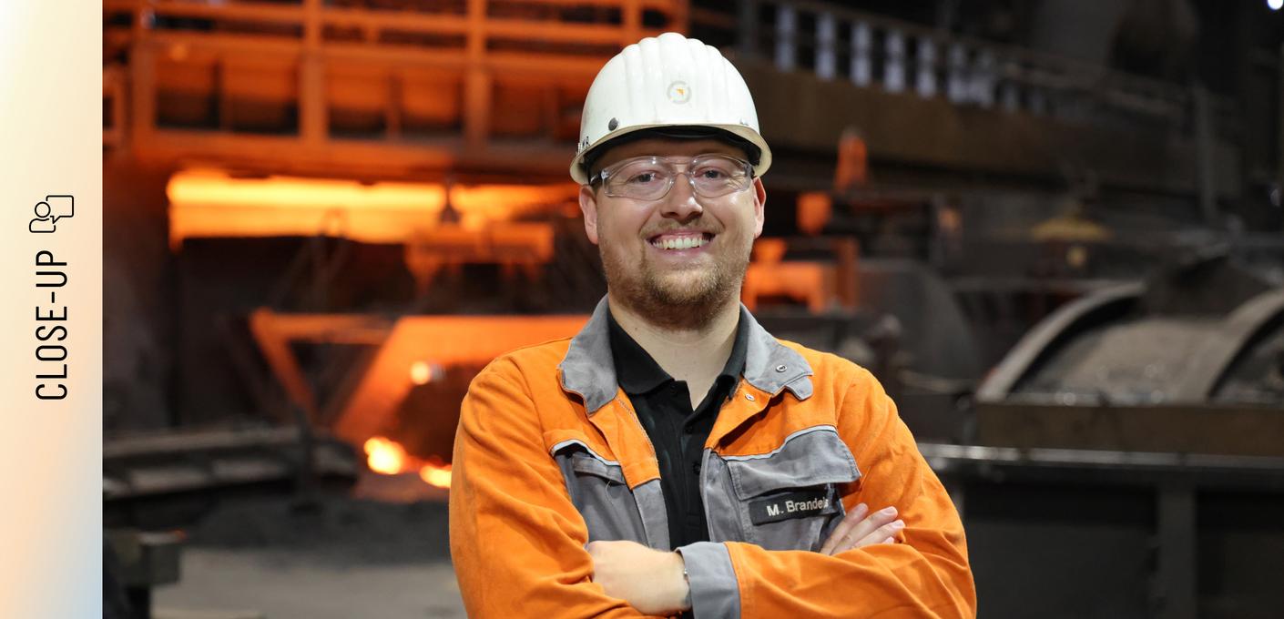 Marvin Brandeis stands in front of a blast furnace with his arms crossed and a smile on his face. He is wearing an orange safety jacket with his name on it, a white hard hat, and safety goggles. In the background, the reddish light of the production facility glows.