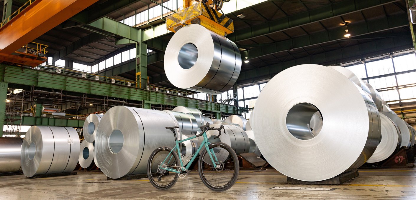 A steel bicycle stands between large, stacked steel coils in an industrial hall, while one coil is being lifted by a crane.