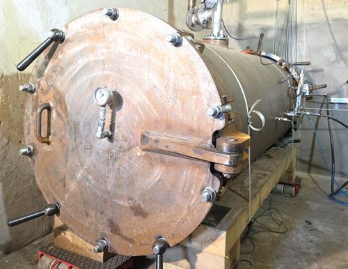 Ein großer, geschlossener zylindrischer Stahlbehälter mit einer runden Öffnung an der Vorderseite steht in einer industriellen Prüfanlage. Der Behälter ist dunkel und massiv, umgeben von Rohrleitungen und technischen Einrichtungen.