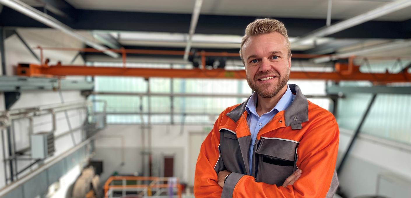 Porträtfoto von Alexander Gering, Gruppenleiter im Bereich Mechanische Prüfung bei der Salzgitter Mannesmann Forschung GmbH in Duisburg. Er trägt orangefarbene Arbeitskleidung und steht mit verschränkten Armen in einer Prüfhalle. Er lächelt freundlich in die Kamera. Im Hintergrund sind Industrieanlagen und Hallenstrukturen zu erkennen.