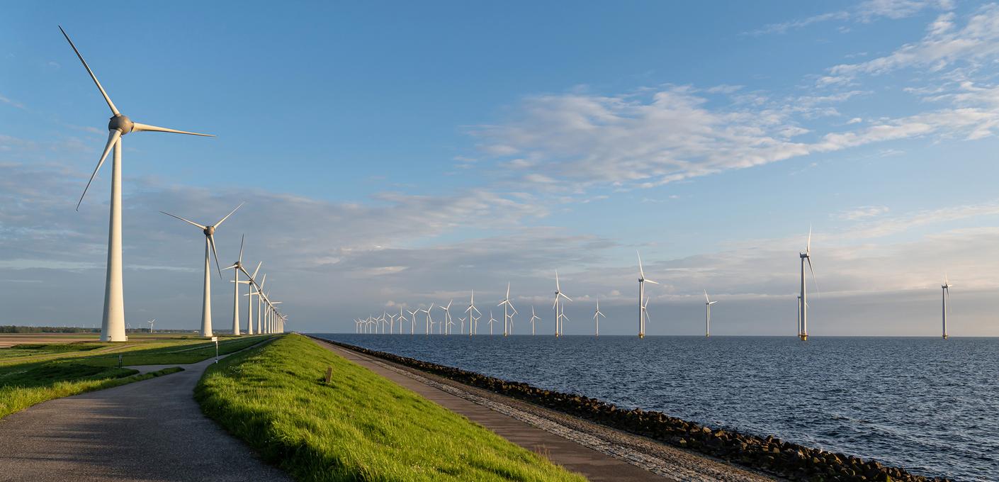 Offshore-Windpark Panoramablick auf einen Offshore-Windpark auf See. Mehrere große Windkraftanlagen ragen aus dem Wasser in einen leicht bewölkten Himmel. Im Vordergrund ist ein grüner Deich zu sehen, dahinter einige weitere Windkraftanlagen an Land.