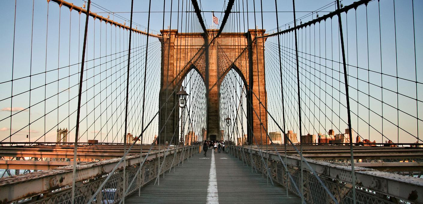 Bild der Brooklyn Bridge. Blick entlang des Fußgängerstegs der Brooklyn Bridge in Richtung eines der steinernen Pylontürme. Stahlseile spannen sich symmetrisch von beiden Seiten zur Mitte. Ein weißer Mittelstreifen führt den Blick auf den gotischen Spitzbogen des sandsteinfarbenen Turms zu, auf dem eine US-Flagge weht. Fußgänger sind in der Ferne erkennbar, das warme Licht der Abendsonne beleuchtet die Szene.