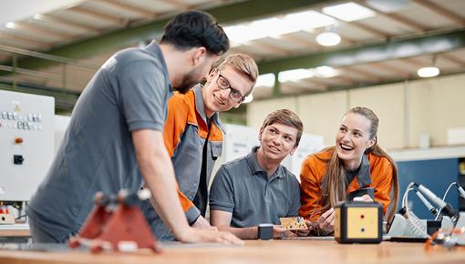 Four young employees in grey and orange workwear stand together at a workbench. They are laughing while examining a workpiece and appear engaged in conversation. Machines and tools are visible in the background.