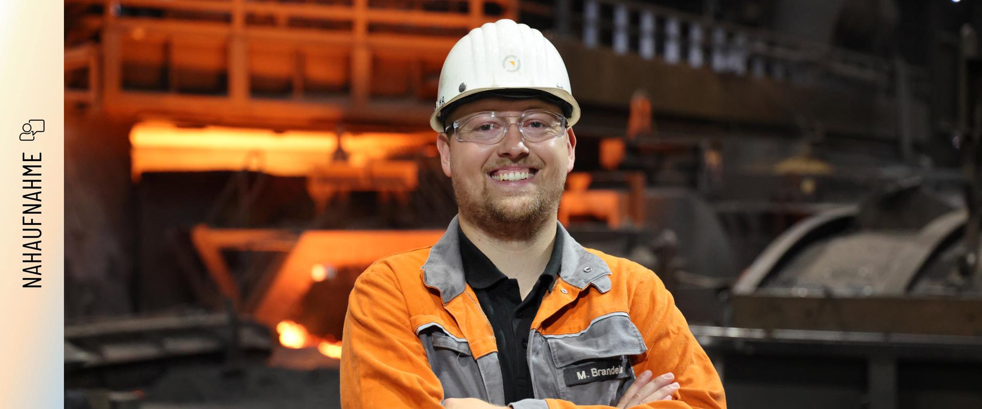 Marvin Brandeis steht mit verschränkten Armen und einem Lächeln vor einem Hochofen. Er trägt eine orangefarbene Arbeitsschutzjacke mit seinem Namenszug, einen weißen Schutzhelm und eine Schutzbrille. Im Hintergrund glüht das rötliche Licht der Produktionsanlage.