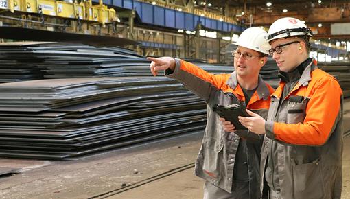 Mitarbeiter stehen vor Stahlblechen in einer Werkshalle Zwei Mitarbeiter in orangefarbener Arbeitskleidung und weißen Schutzhelmen stehen vor gestapelten Stahlblechen in einer Werkshalle. Einer zeigt mit ausgestrecktem Arm in die Ferne, der andere hält ein Tablet und blickt in dieselbe Richtung.