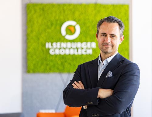Thorsten Gintaut, Geschäftsführer bei der Ilsenburg Grobblech GmbH, im Porträt Porträtfoto von Thorsten Gintaut, Geschäftsführer bei der Ilsenburg Grobblech GmbH. Er trägt ein dunkles Hemd und steht mit verschränkten Armen vor einer Wand mit dem Logo der Ilsenburger Grobblech GmbH. Er lächelt selbstbewusst in die Kamera.