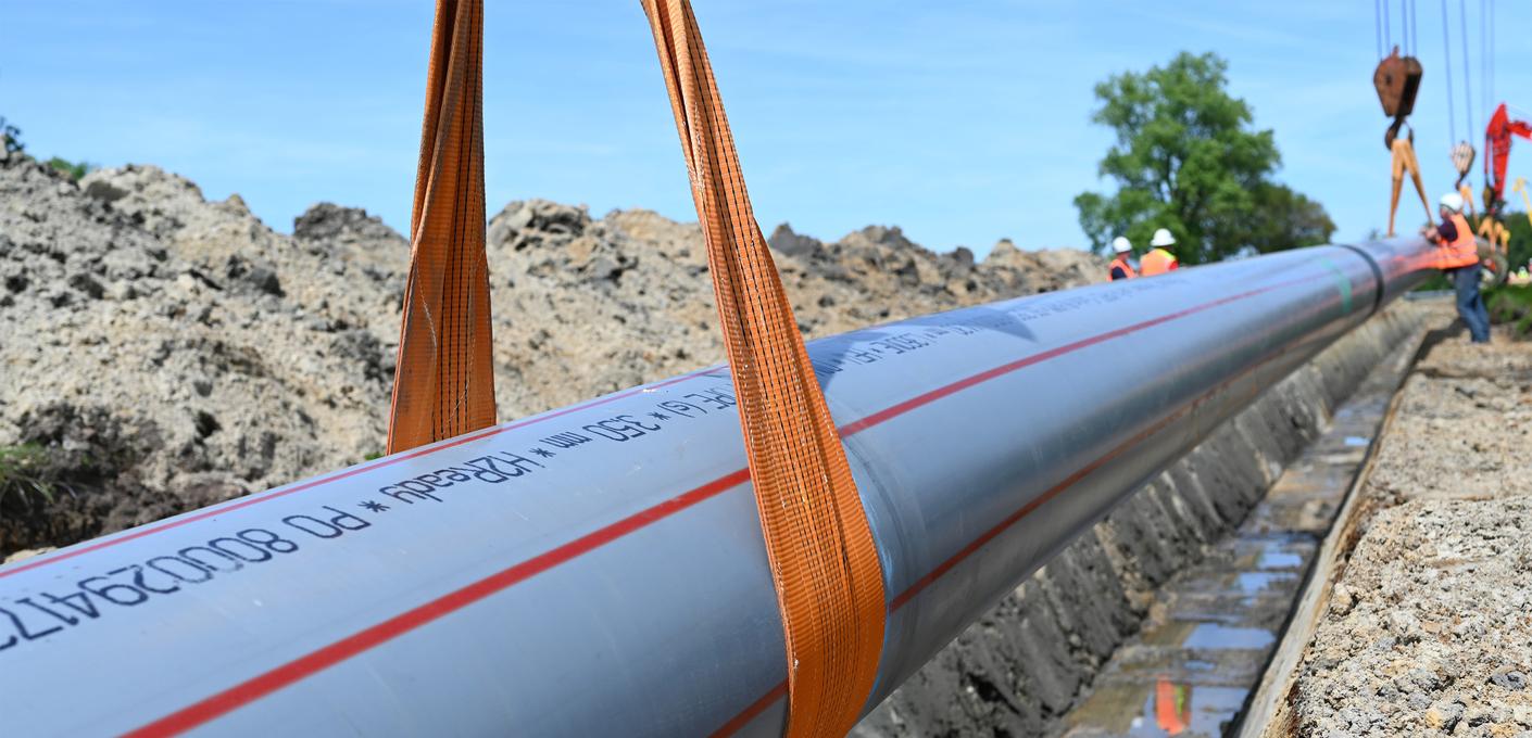 The H2Ready pipe is being lowered into an open trench at a construction site with the help of lifting straps and cranes. The H2Ready pipe is being lowered into an open trench at a construction site with the help of lifting straps and cranes.