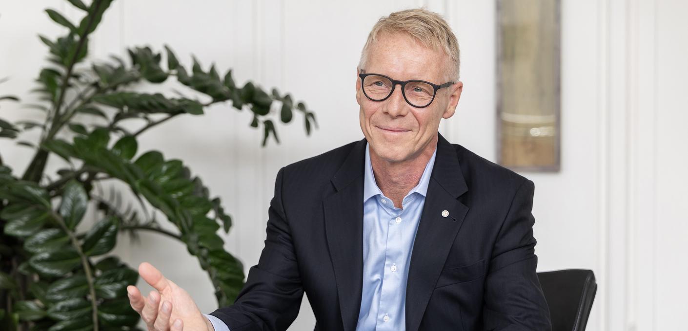 Portrait photo of Benedikt Ritterbach, Managing Director of Salzgitter Mannesmann Forschung GmbH (SZMF). He wears glasses, a white shirt and a dark blazer and looks friendly into the camera. Blurred plants and a bright interior are visible in the background.